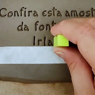 Detalhe: a mão encosta a letra na fita crepe, que serve de régua sobre a argila